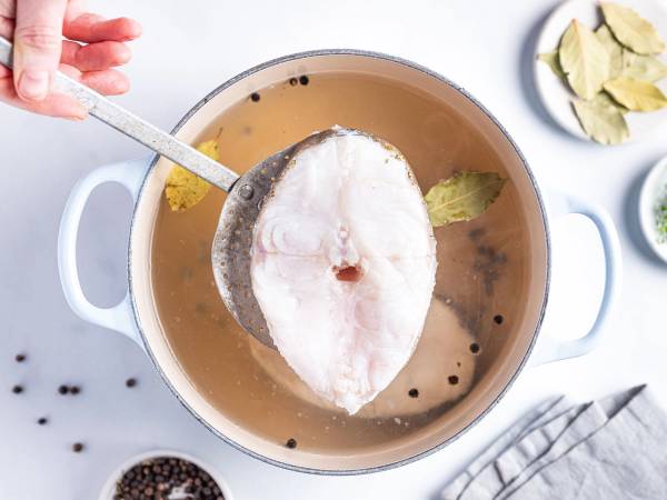 En person bruker en skje til å fjerne skreien fra en kjele med kraft eller suppe, med flere krydder og urter rundt på bordet.