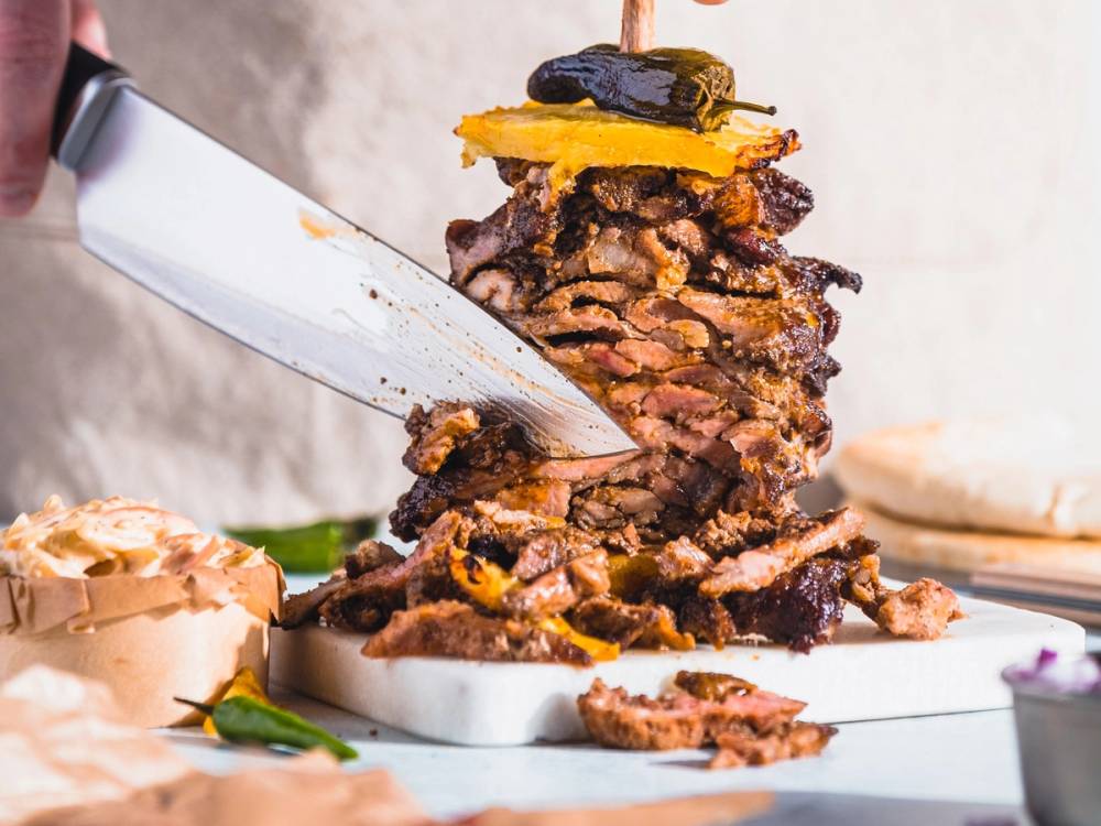 En stor shawarma med kjøtt, løk og ananas på en tallerken, med en kniv som skjærer i den.