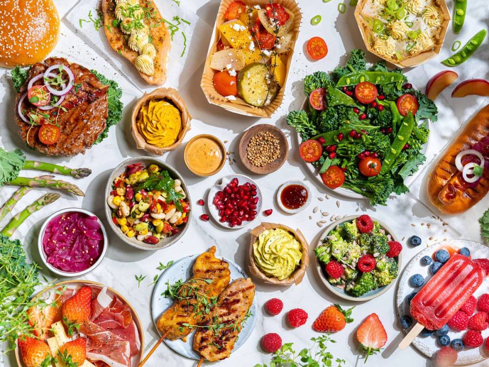 Et utvalg av sommermat med salater, frukt, grillmat og is på et dekket bord.