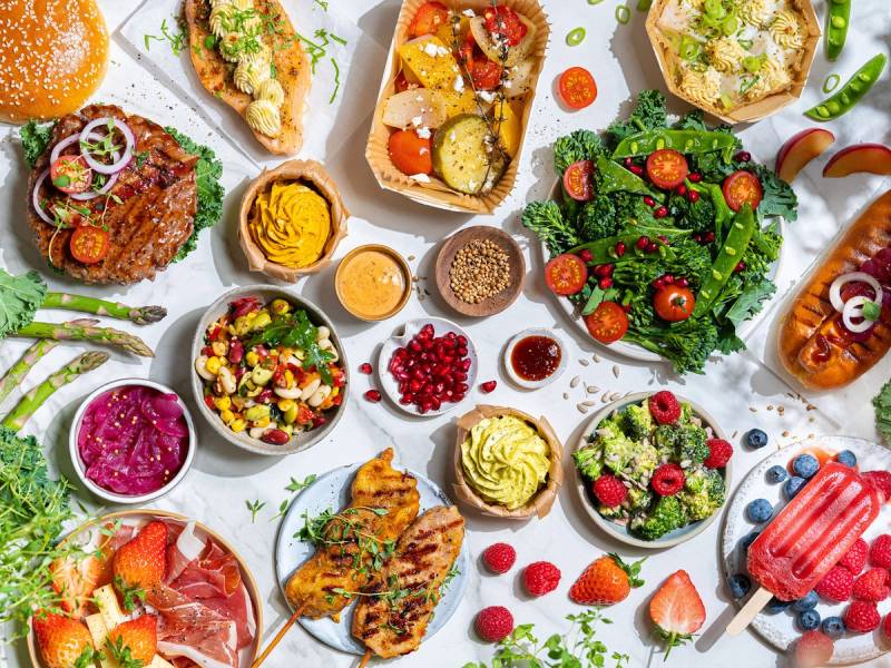 Et utvalg av sommermat med salater, frukt, grillmat og is på et dekket bord.