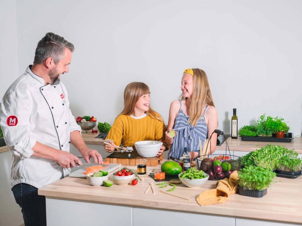 To barn og en kok i et kjøkken med friske grønnsaker og matlagingsutstyr, ser ut til å lage mat sammen.