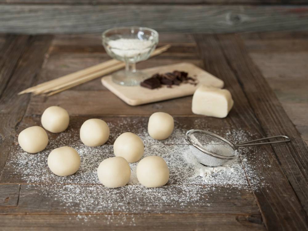 Hjemmelaget marsipansnøballer på et bord med mel og marsipan, samt et par pinner og en liten skål med melis eller mel, klar til å formes.