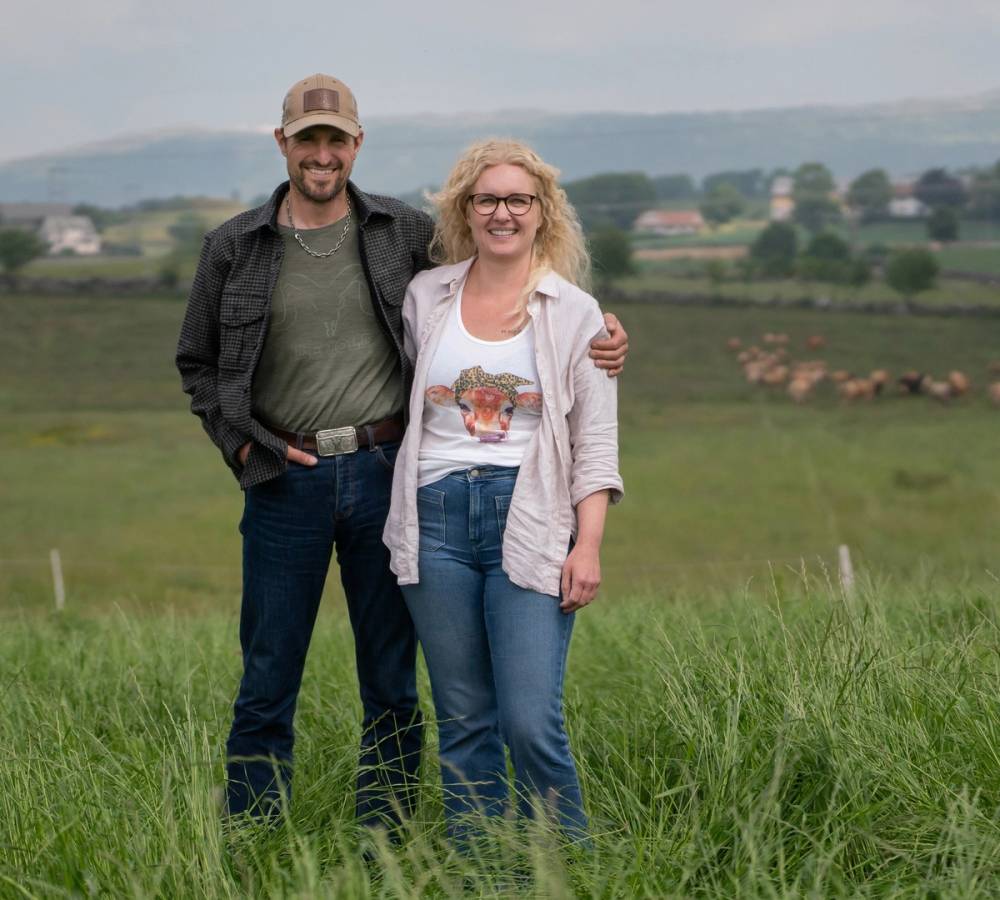 En mann og en kvinne står sammen i en grønn eng med landskap og hus i bakgrunnen, og de har armene rundt hverandre.