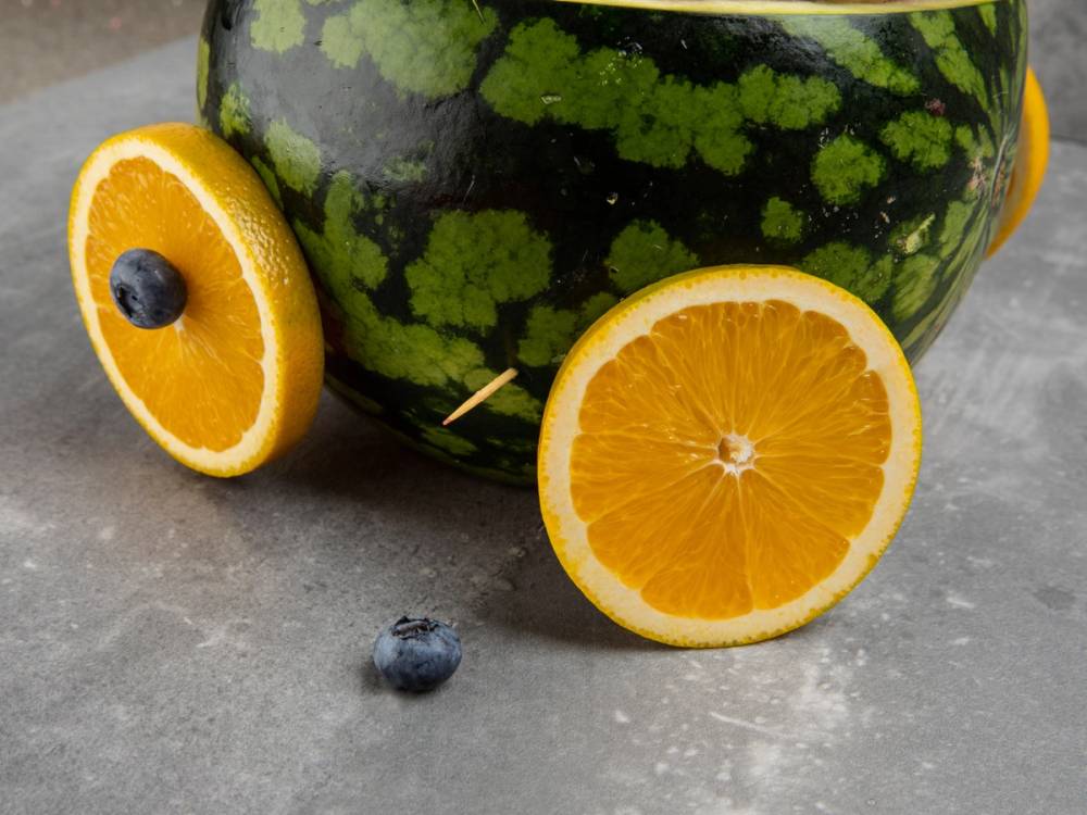 En vannmelon med skiver av appelsin og en blåbær foran på en grå overflate, sannsynligvis som del av en babyshower-dekorasjon.