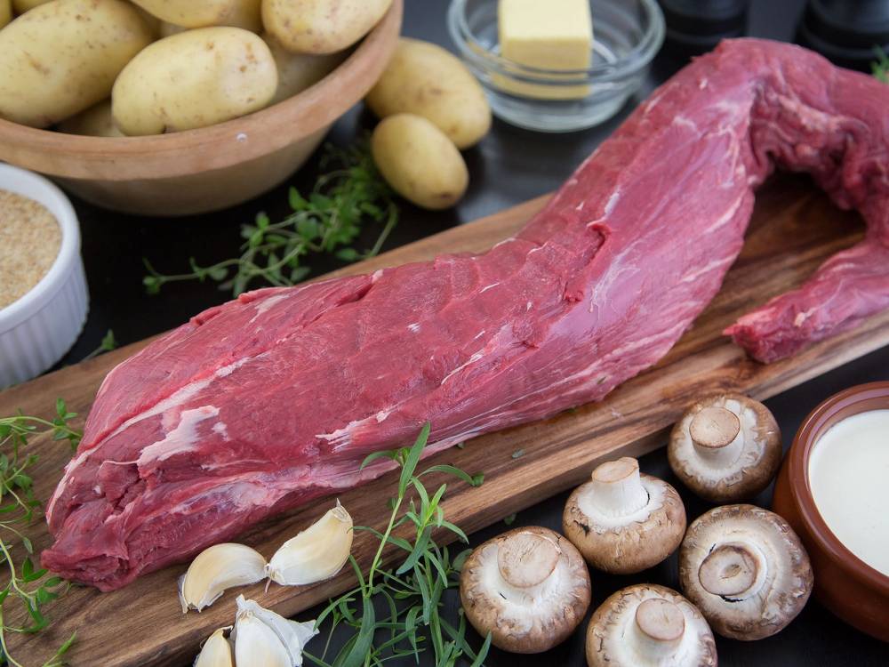 Utsnitt av en rå indrefilet av okse på skjærebrett omgitt av sopp, hvitløk, poteter og krydderurter.