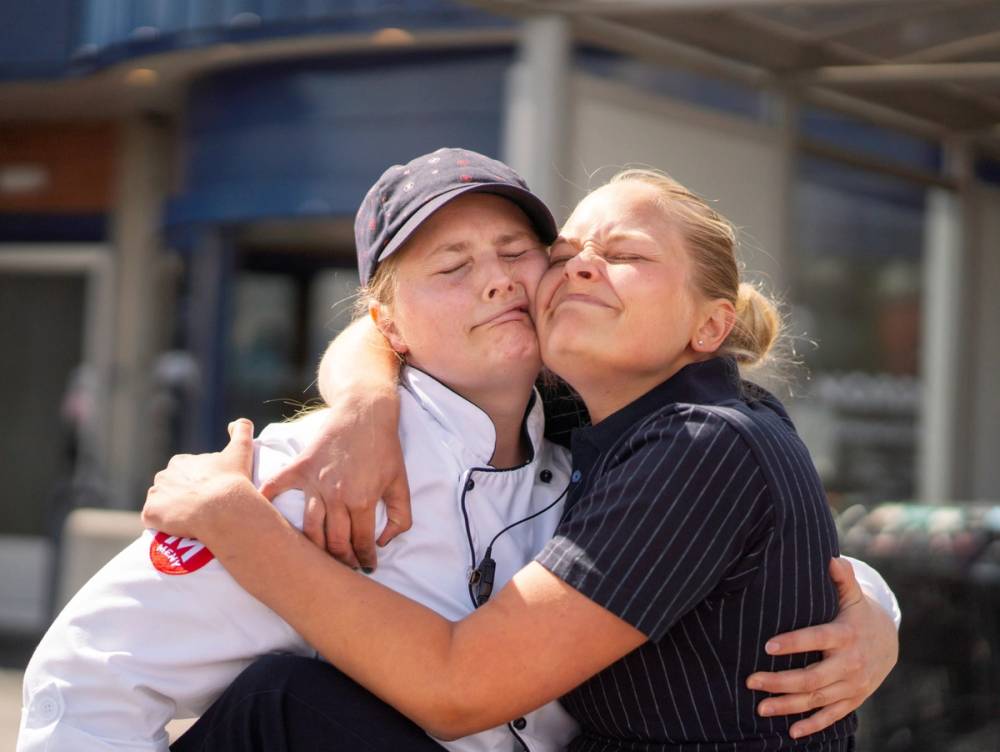To personer omfavner hverandre i en klem utenfor en butikk, med en person i kokkeuniform og en annen i arbeidsklær.