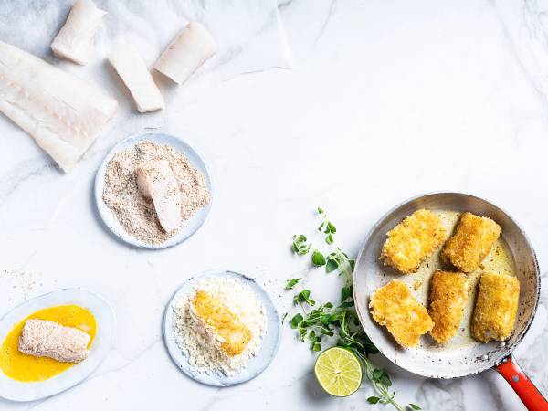Fiskepinner på en tallerken med sitron og urter, omgitt av brødsmuler og en skål med mel, på et hvitt bord.