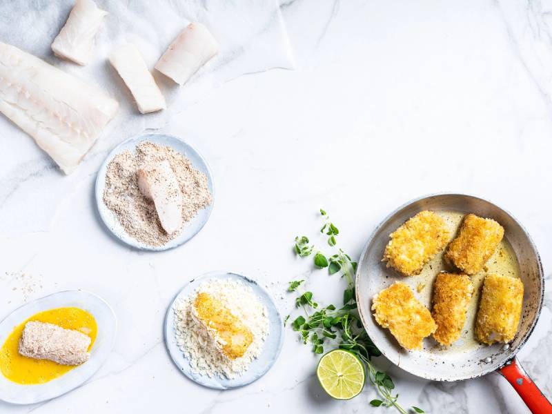 Fiskepinner på en tallerken med sitron og urter, omgitt av brødsmuler og en skål med mel, på et hvitt bord.