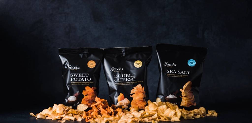 Tre poser med snacks, inkludert søtpotet, ost og sjøsalt, plassert foran en mørk bakgrunn, med noen knuste snacks rundt.