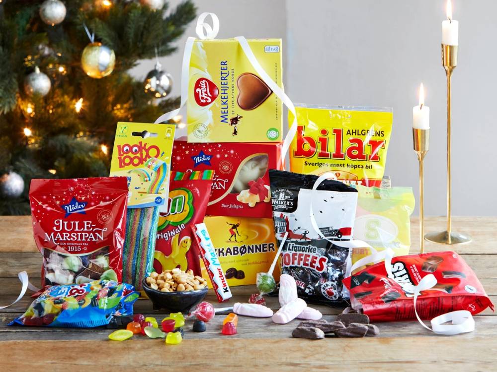 Et utvalg av julegodterier og sjokolade på et bord foran en julegran med lys, med godterier som marsipan, sjokoladeplater og små godterier, og en julekalender i bakgrunnen.