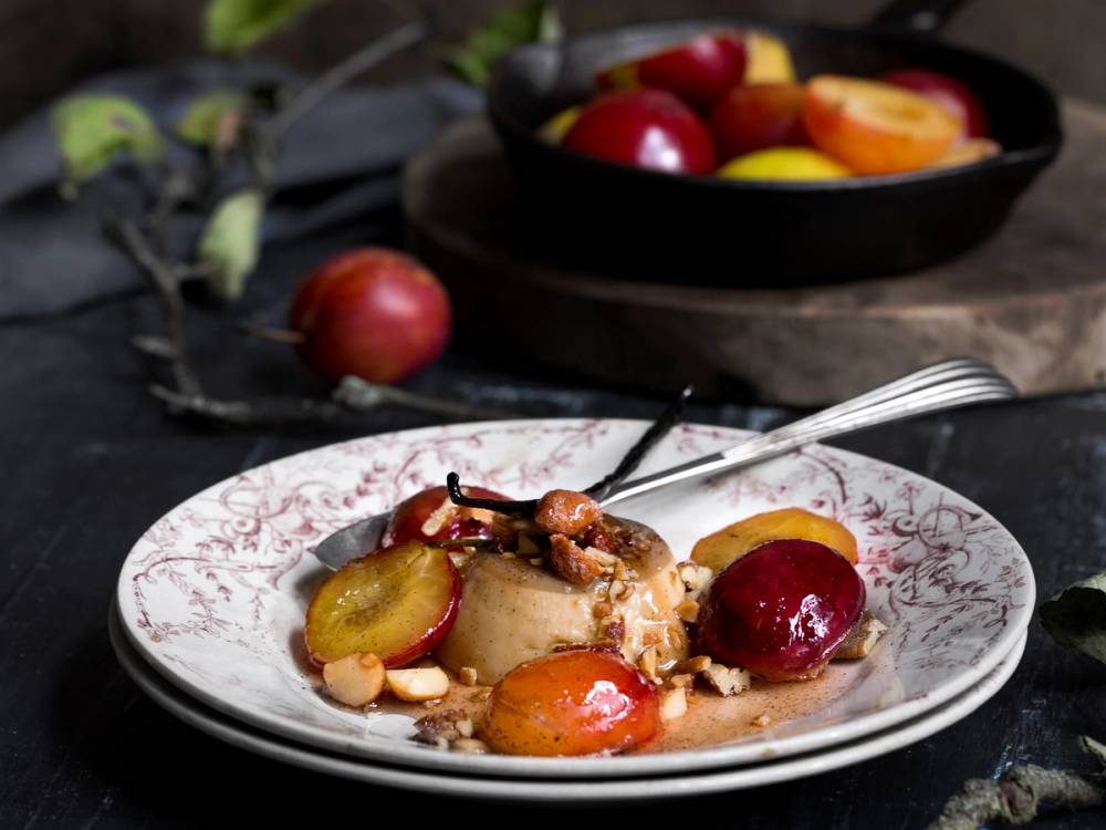 Panna cotta med kaffe og plommer servert på en tallerken med en skje ved siden av, med en skål med flere plommer i bakgrunnen.