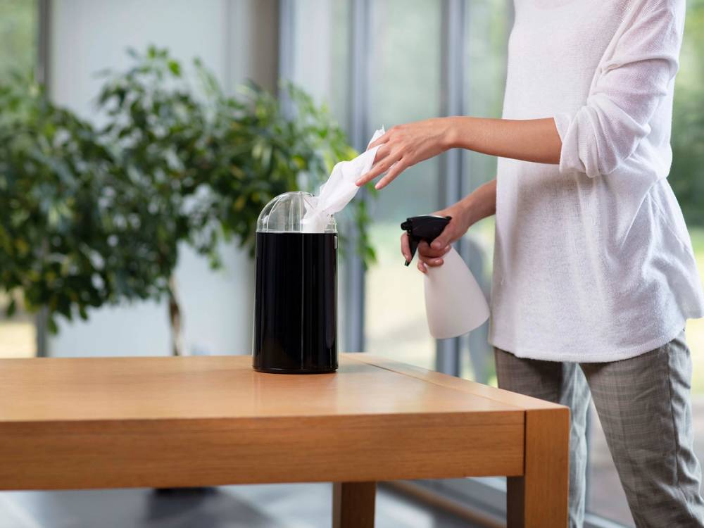 Person kaster brukt tørkepapir i en svart søppelbøtte på et bord i et rom med store vinduer og grønne planter i bakgrunnen.