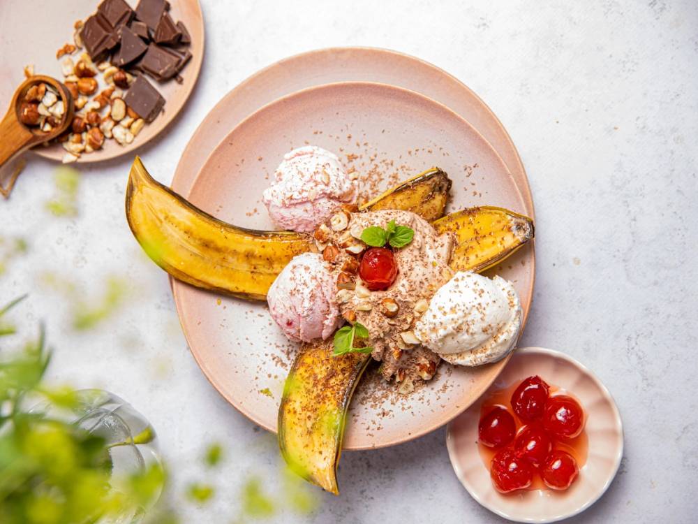 Banansplitt med sjokoladekrem og jordbær på en rosa tallerken, omgitt av små skåler med sjokolade og kirsebær.