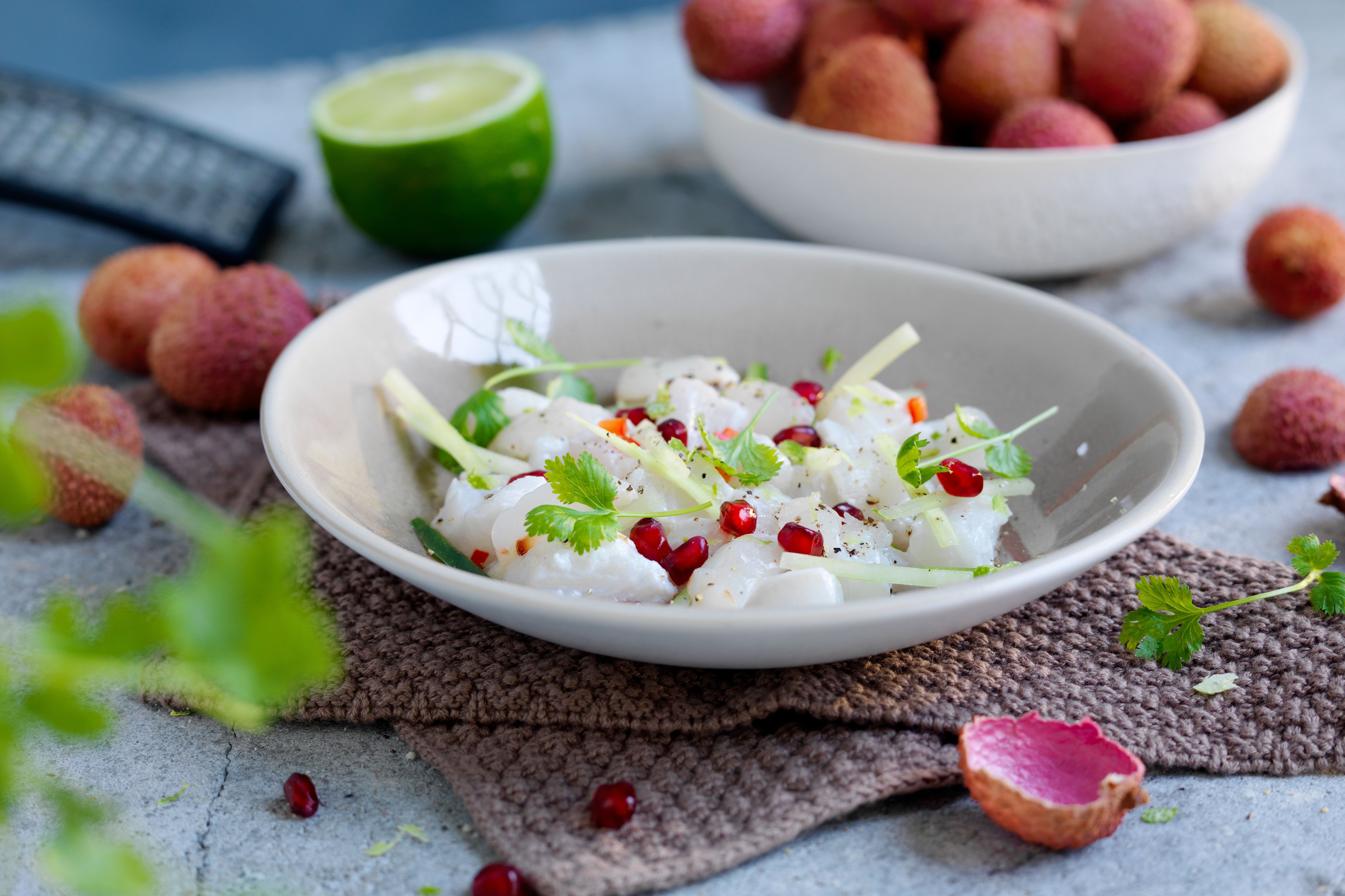 En skål med ceviche med litchi, pyntet med urter og små røde bær, omgitt av lime og lyse frukt på et bord.