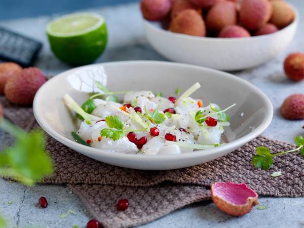 En skål med ceviche med litchi, pyntet med urter og små røde bær, omgitt av lime og lyse frukt på et bord.