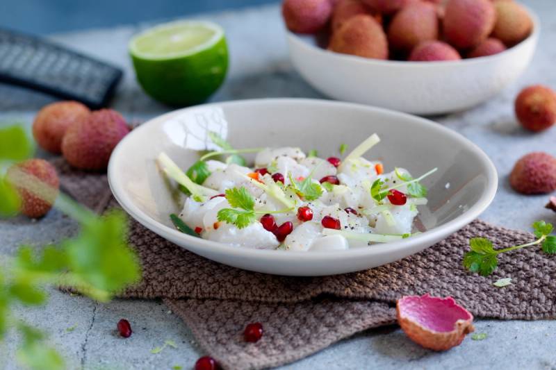 En skål med ceviche med litchi, pyntet med urter og små røde bær, omgitt av lime og lyse frukt på et bord.