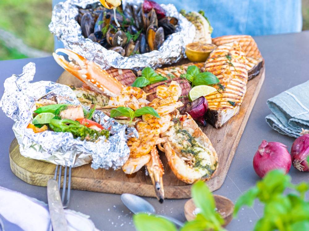 Grillmat med skalldyr, laks og grønnsaker på et serveringsfat, omgitt av tilbehør og sitronbåter.