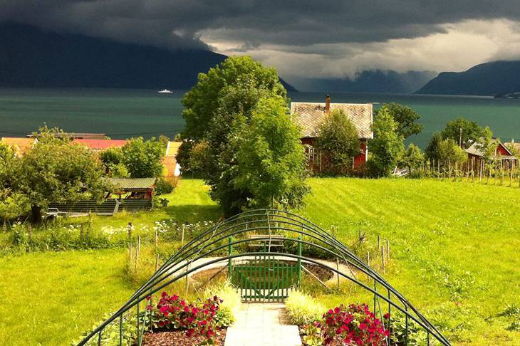 Et grønt landskap med en stor tre, en hage med blomster og en liten bygning, med fjell og vann i bakgrunnen under en overskyet himmel.