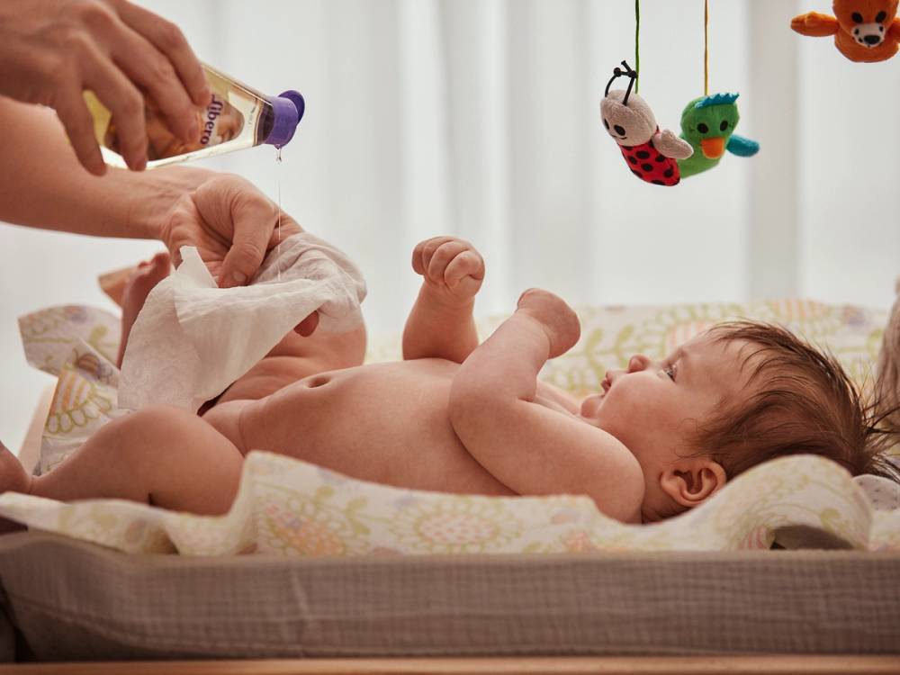 En baby ligger i en seng og blir skiftet på med bleie, mens en leke med flere farger henger over sengen.