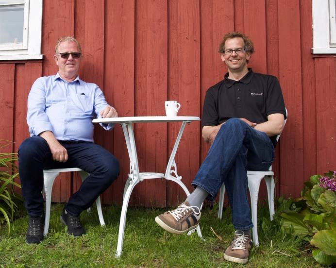 To personer sitter på verandaen med en liten hvit bord mellom seg, foran en rød vegg, og det er en kopp på bordet.