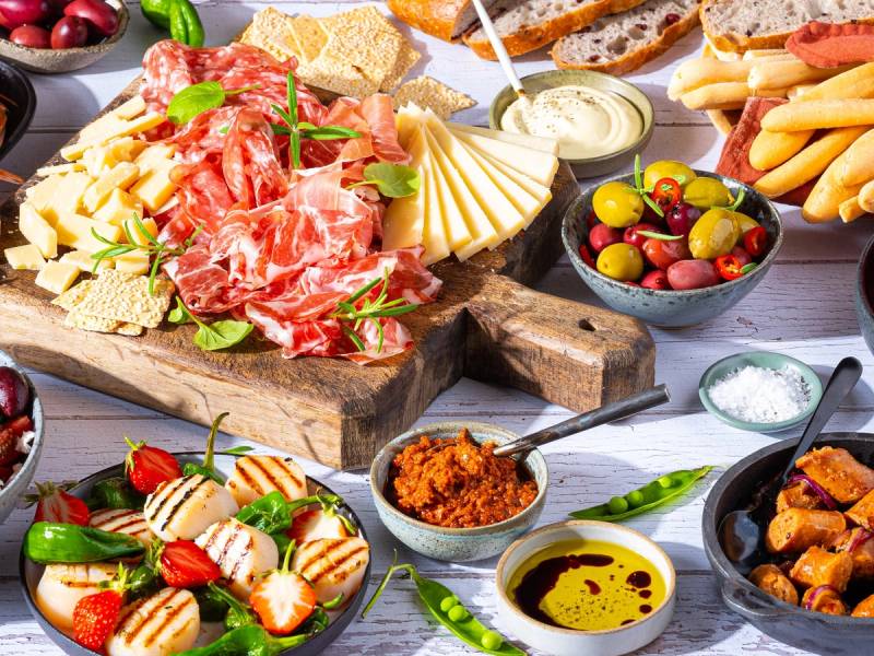 Et utvalg av middelhavsretter med ost, skinke, oliven, brød og ulike dipper på et bord.