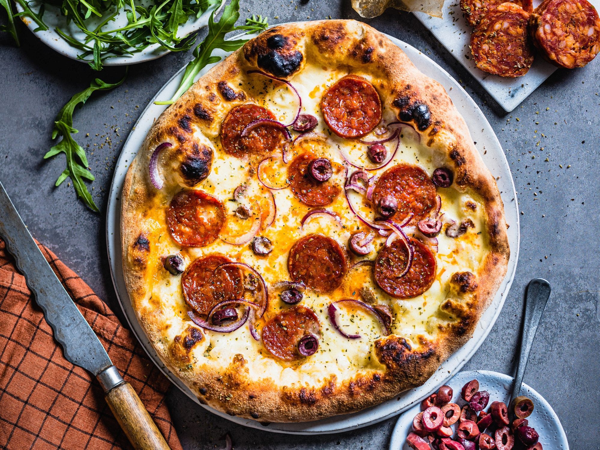 En pizza med pepperoni, ost og løk på en svart stekeplate, omgitt av grønne urter og små skåler med tilbehør.