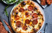 En pizza med pepperoni, ost og løk på en svart stekeplate, omgitt av grønne urter og små skåler med tilbehør.