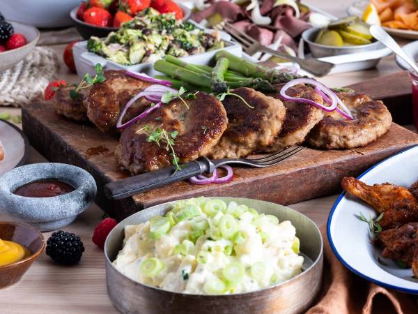 Et koldtbord med ulike typer kjøtt, grønnsaker, potetsalat og frukt, med en person som legger til mer mat i bakgrunnen.