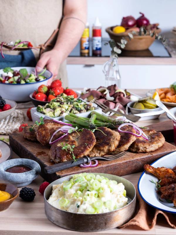 Et koldtbord med ulike typer kjøtt, grønnsaker, potetsalat og frukt, med en person som legger til mer mat i bakgrunnen.