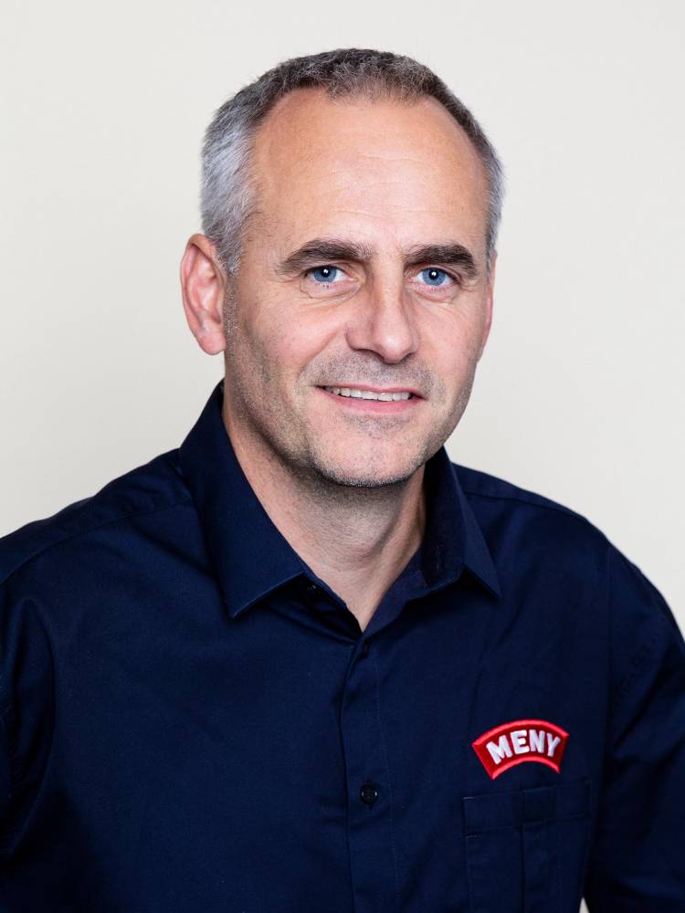 En mann med grått hår iført en mørkeblå skjorte med MENY-logo, ansiktet er dekket av en rosa blokk.