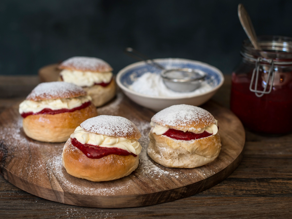 Fire fastelavnsboller med vaniljekrem og et lag med jordbærsyltetøy på en rund treplate, omgitt av en skål med melis og et glass med rød syltetøy.