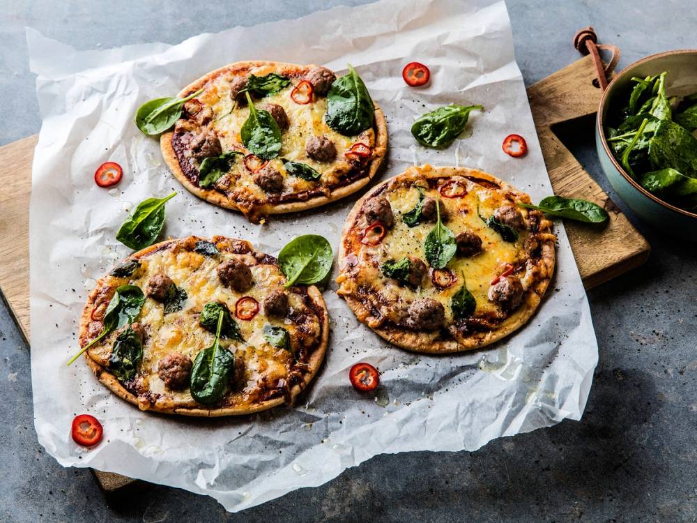 Tre pizzaer med ost, basilikum og cherrytomater på bakepapir, med en liten skål med salat i bakgrunnen.