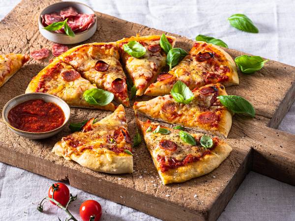 Pizzaskive med flere pizzaer med ost, skinke og basilikum på en treplate, omgitt av tomater og basilikumblader.