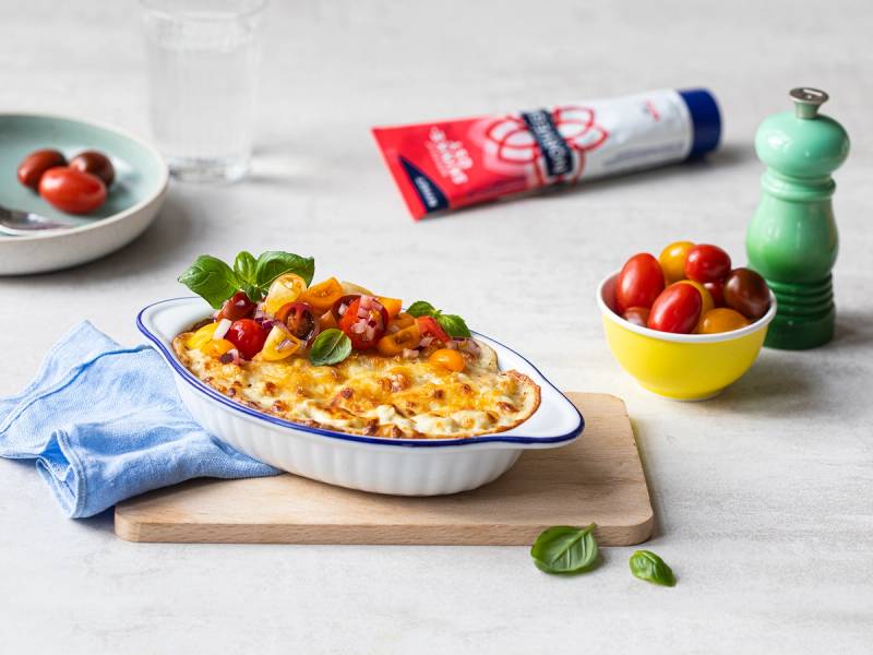 En porsjon med smøreost på en tallerken med tomater og basilikum, omgitt av en tube med Tine smøreost og en grønn pepperkvern på et kjøkkenbord.