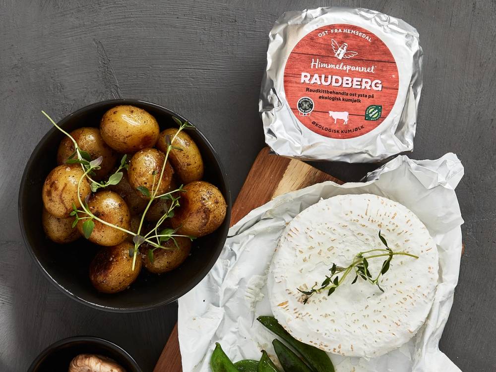 Et bilde av en pakke med Raudberg-ost, en tallerken med poteter og en tallerken med ost og urter på et bord.