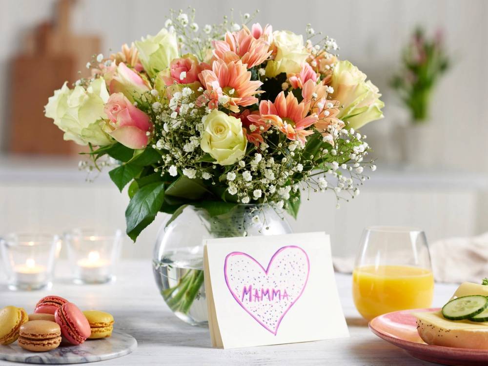 En bukett med rosa og hvite blomster, inkludert roser og gerbera, på et bord med en kort som sier 'Mamma' og små kaker og juice rundt.