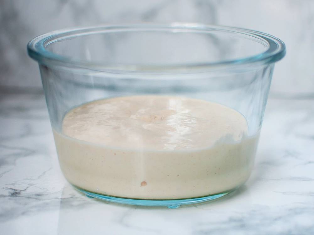 En glassbolle med en hvit, fermentert deig eller poolish som har hevet, plassert på en hvit bakgrunn.