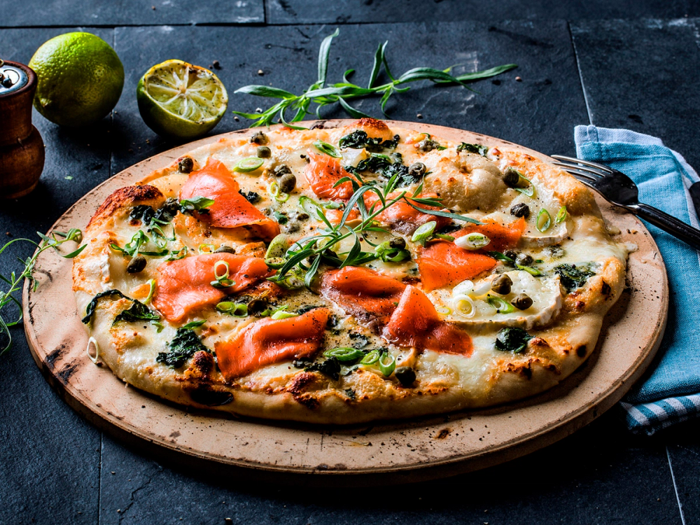 Hvit pizza med røkelaks, spinat og grønne urter på en treplate.