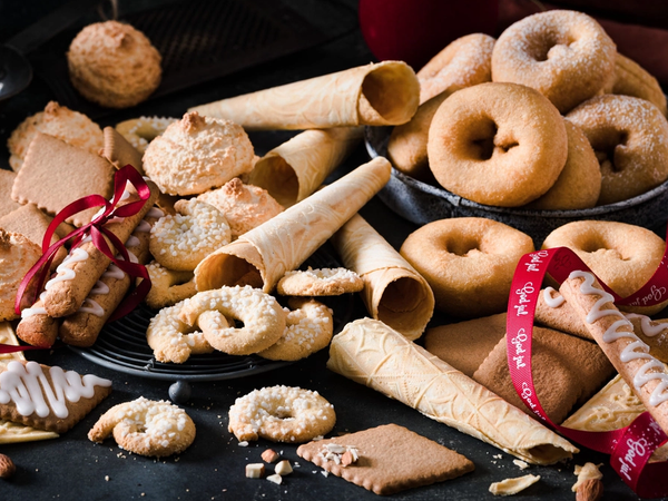 Et utvalg av tradisjonelle julekaker på et svart fat, inkludert krumkaker, sandkaker og småkaker med melis, omgitt av julekuler og en rød julebånd.