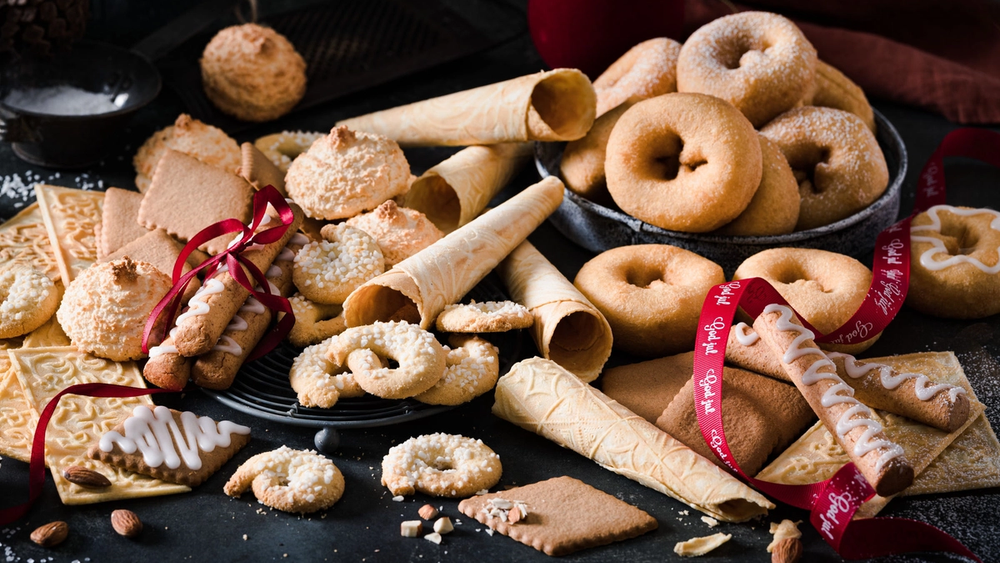 Et utvalg av tradisjonelle julekaker på et svart fat, inkludert krumkaker, sandkaker og småkaker med melis, omgitt av julekuler og en rød julebånd.