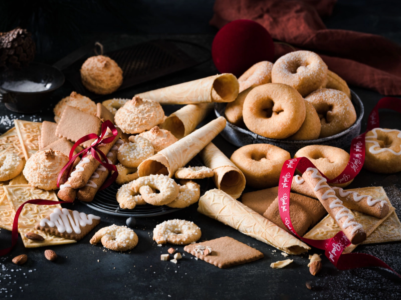 Et utvalg av tradisjonelle julekaker på et svart fat, inkludert krumkaker, sandkaker og småkaker med melis, omgitt av julekuler og en rød julebånd.