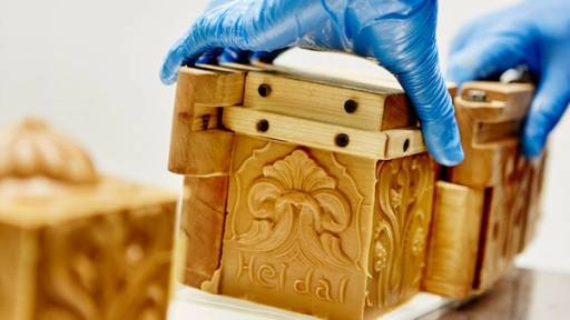 Hender i blå hansker som håndterer en treboks med utskjæringer og dekorasjoner, sannsynligvis i en produksjonsprosess for Heidal-ysteri.