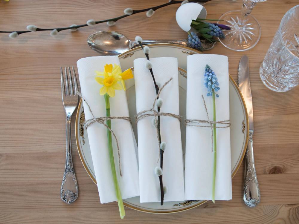 På et påskebord ligger hvite servietter med små påskeliljer og blå blomster som dekorasjon, omgitt av sølvbestikk og glass.