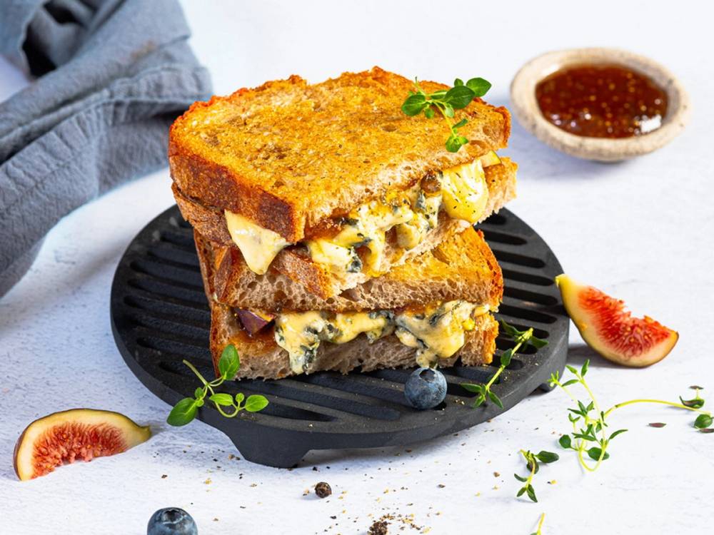 To toast med Selbu Blå ost på en svart tallerken, omgitt av ferske blåbær, fiken og en liten skål med syltetøy, på et hvitt bord.