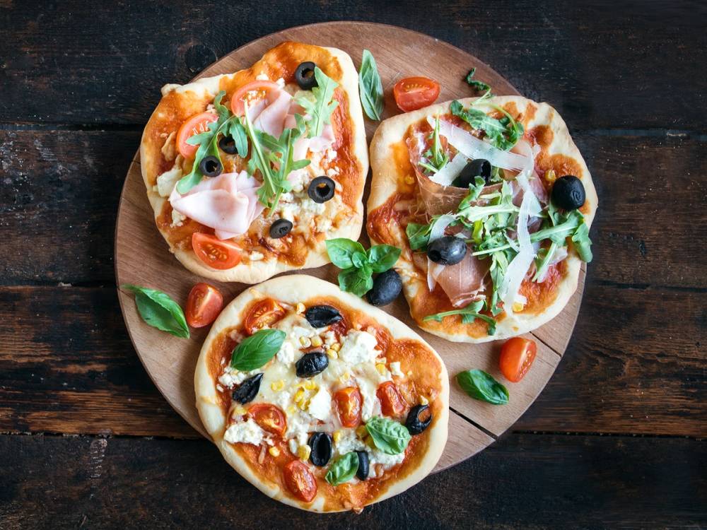 Tre små pizzaer med ost, skinke, oliven og grønne urter på en rund treplate