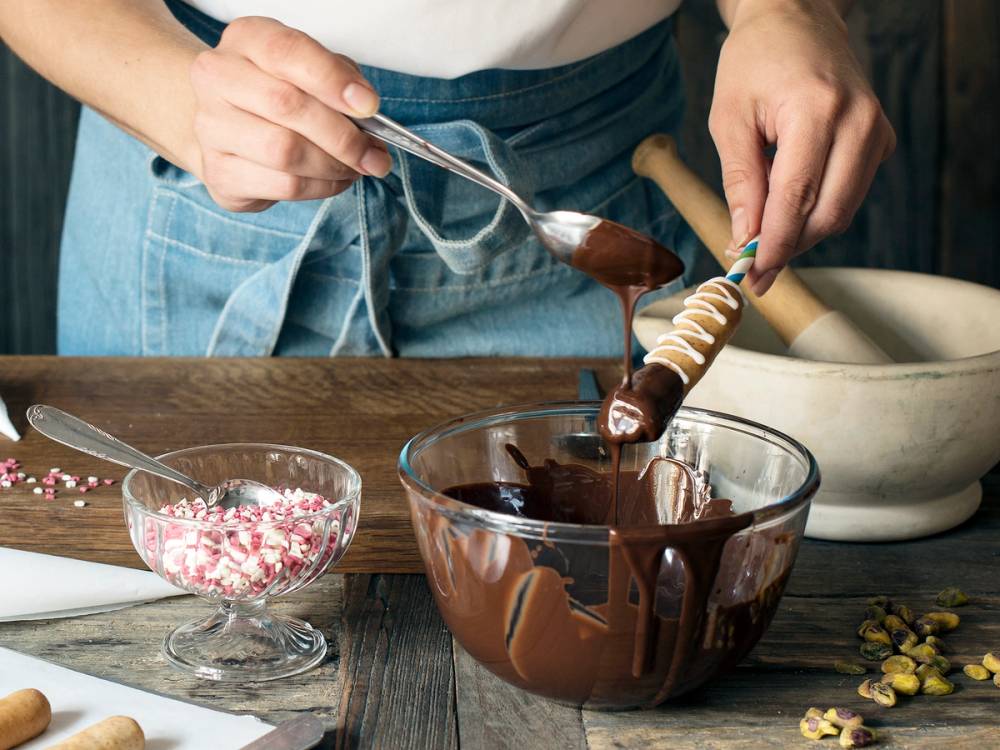 Person som dekorerer en kransekakestang med sjokolade og strøssel, med en bolle med smeltet sjokolade foran seg.
