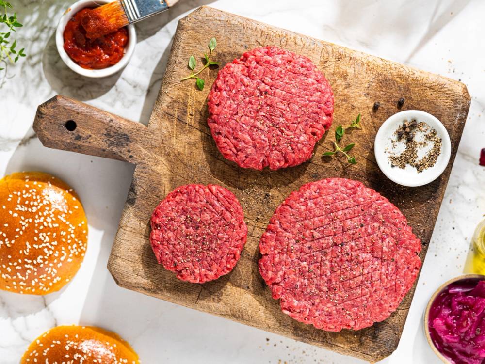 Tre rå hamburgere av kjøttdeig på en skjærebrett med små skåler av krydder og saus rundt.