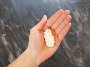 En person holder en hånd med en enkelt nigiri-sushibit, bestående av ris og fisk, mot en mørk bakgrunn.