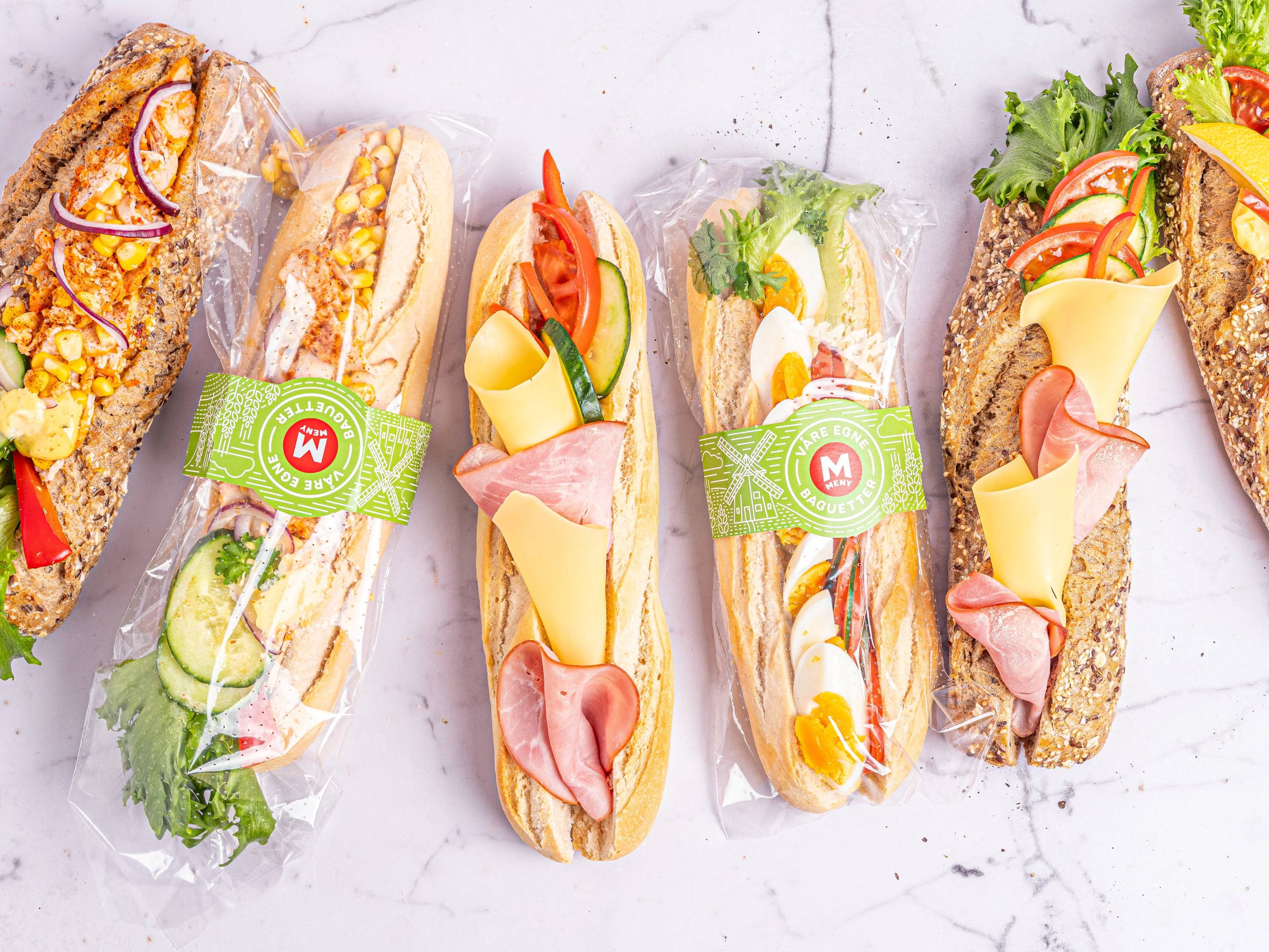 Fire baguetter med ulike pålegg og grønnsaker på et hvitt underlag