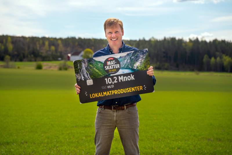 Person holder opp en stor sjekk med teksten '10,2 Mnok' og 'Lokalmatprodusenten' på en grønn åker under en blå himmel.